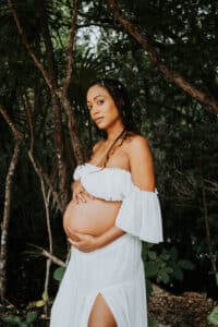 Pregnant woman in flowing dress standing in the lush jungle of Tulum, cradling her belly during a serene maternity photo shoot