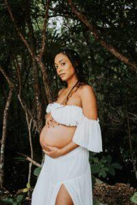 Pregnant woman in flowing dress standing in the lush jungle of Tulum, cradling her belly during a serene maternity photo shoot