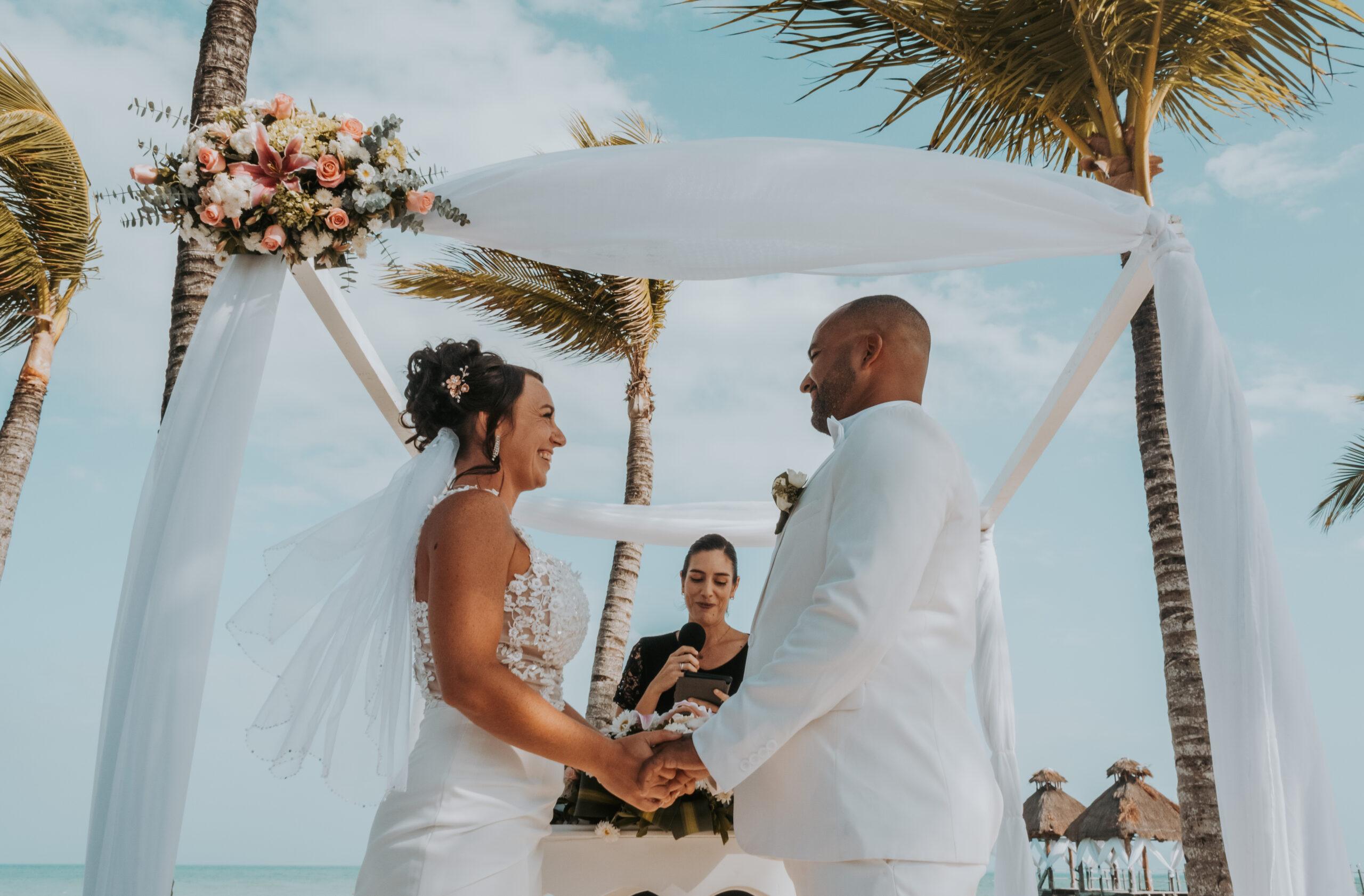 Pareja dándose el ‘sí’ durante una ceremonia de boda en la playa, al atardecer, con el mar de fondo y decoración floral.