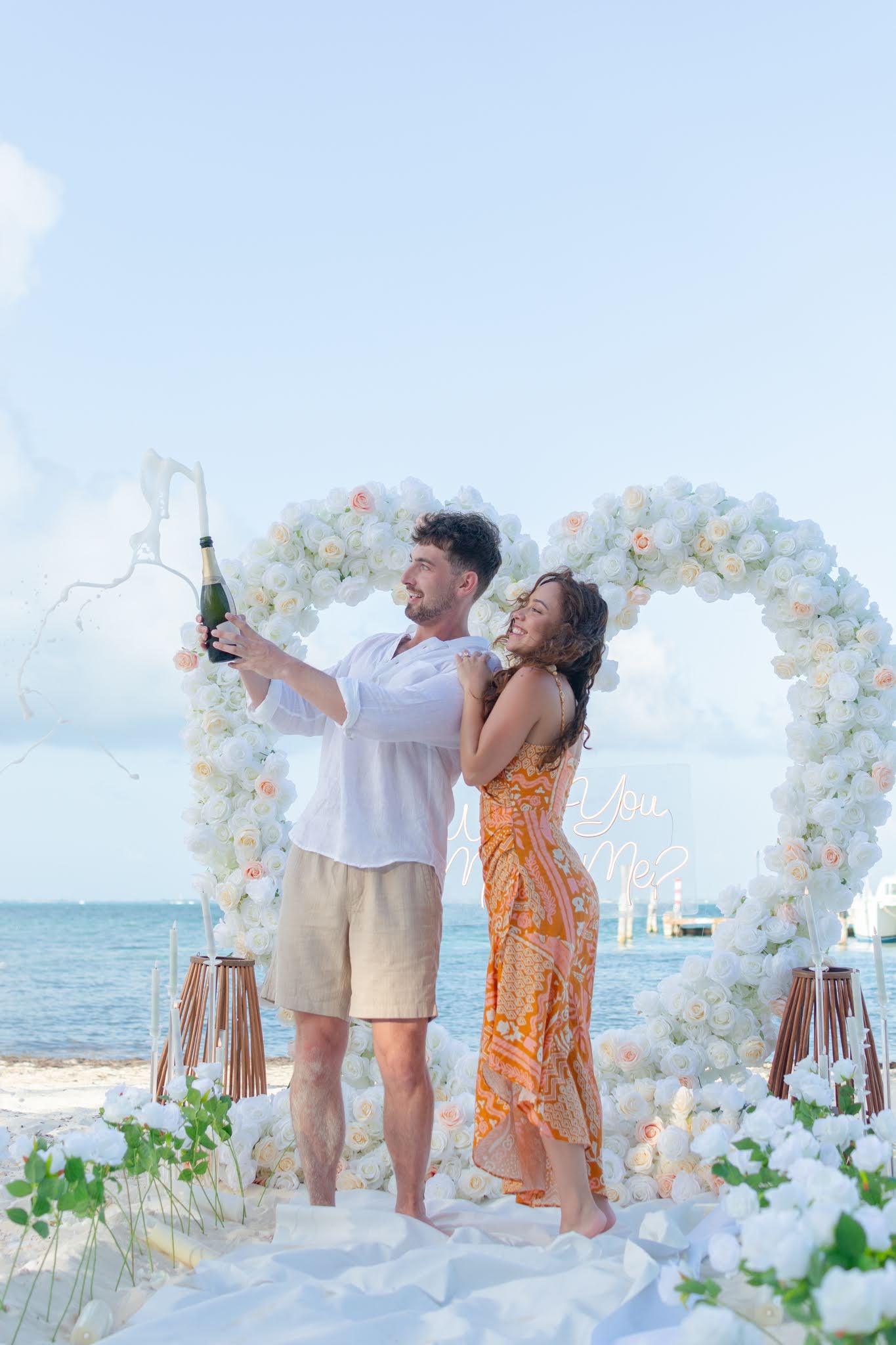 Couple toasting with champagne in front of a heart made of red flowers during a romantic beach photo session