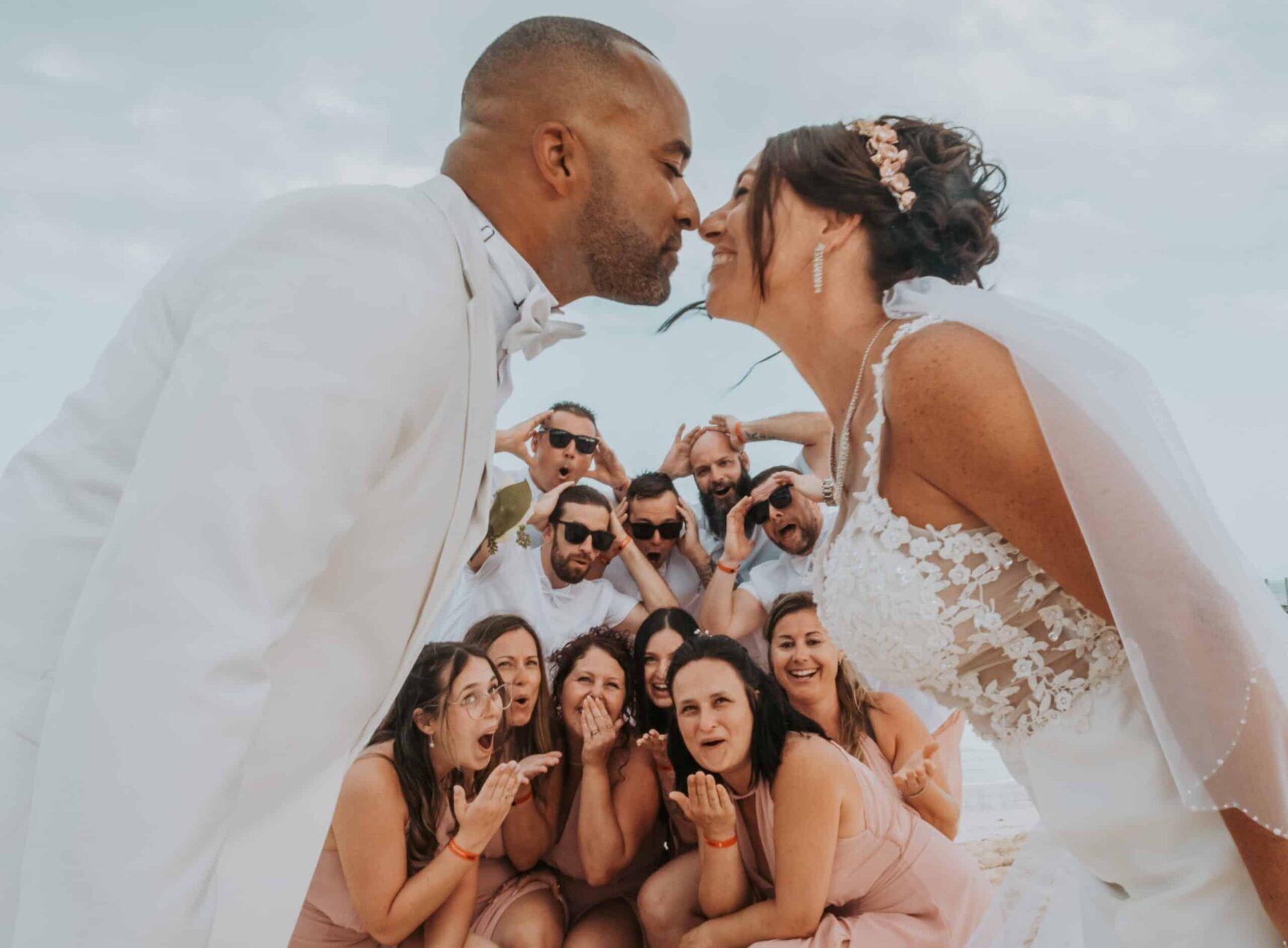 Bride and groom leaning in for a kiss with their bridal party cheering behind them – a playful wedding portrait photo perfect for wedding photos framed.